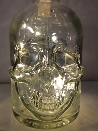 A clear glass bottle shaped like a skull dominates the image. The skull's features, including eye sockets, nose cavity, and teeth, are prominently molded into the glass. Light refracts through the glass, creating bright highlights and darker shadows that define the skull's form. The bottle sits on a softly lit surface, adding to the overall luminous effect. The background is a muted gray, further emphasizing the bottle's transparency and intricate design.