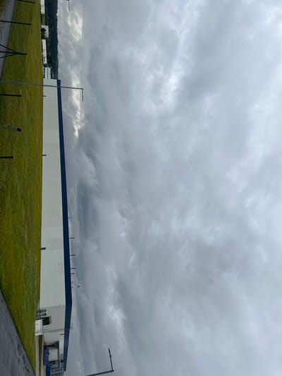 An exterior, eye-level, medium shot captures the side of a long, white industrial building with a dark blue trim. The building's facade is mostly uninterrupted, but it features several loading docks, windows, and external ladders. A section of a chain-link fence borders a lush green field to the left of the building, and a small red and white circular sign is visible on the fence. The ground in front of the building appears wet and paved. The sky is overcast with heavy grey clouds, obscuring any clear view of the horizon or distinguishing weather patterns. In the background, a line of trees is visible, and the faint outlines of power lines and towers can be discerned. There are no people or animals visible in the frame. The image appears to be taken in the city of Ostrov u Stříbra, Czechia, based on the provided context.