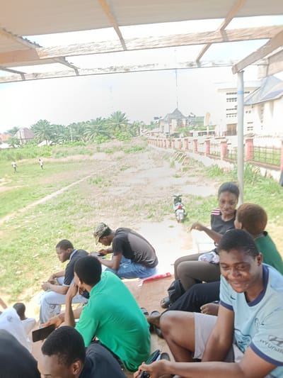 In Ukpor, Nigeria, a group of young people are seated on a tiered structure, likely bleachers, under a partially constructed or weathered roof. The background shows a grassy field with scattered palm trees and buildings, suggesting a semi-rural or developing urban area. In the foreground, several individuals are engaged with their mobile phones. One person in a light blue t-shirt smiles towards the camera while holding a dark object, possibly a game controller or another phone. Others are looking down at their devices, some with earbuds in. A motorcycle is parked in the mid-ground. The lighting indicates it is daytime with bright sunlight casting shadows.