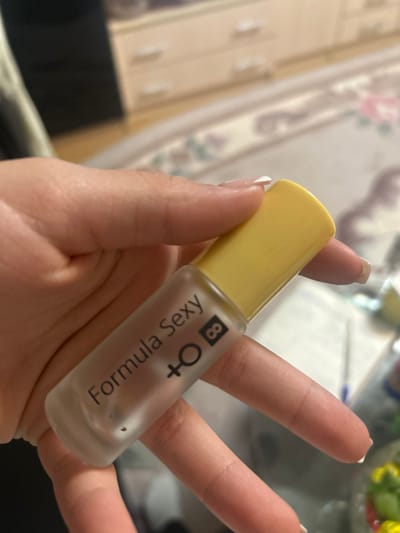 A light-skinned hand, displaying manicured nails with white tips, holds a small, frosted glass bottle vertically. The bottle features a bright yellow cap and bears the black inscription "Formula Sexy + ♀ 8" on its side. The background, out of focus, depicts an indoor setting in Dmitriyevskaya, Russia, characterized by a light-colored rug with a floral pattern, a beige dresser with white handles, and a blue pen visible on a surface to the right.