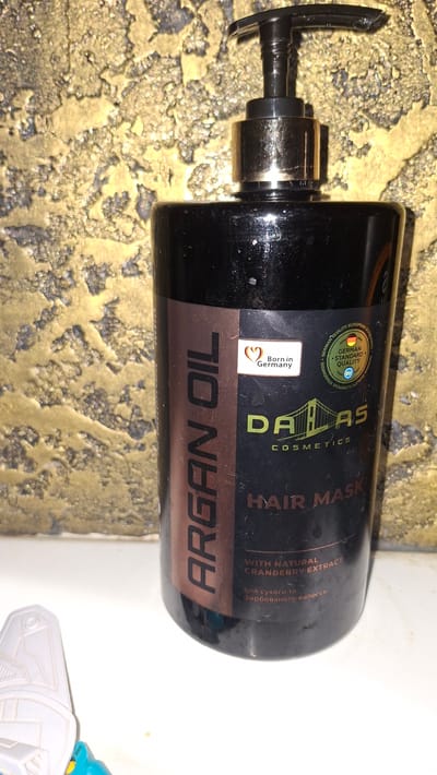 The image shows a black bottle of "DALAS COSMETICS HAIR MASK" with "ARGAN OIL" and "WITH NATURAL CRANBERRY EXTRACT" printed on the label. There are also indications of "Born in Germany" and "GERMAN STANDARD QUALITY". The bottle has a pump dispenser. The background is a textured wall with a goldish-yellow hue. A white and blue object is partially visible in the lower left corner, appearing to be a toy. The image is taken indoors. There is no information or visual evidence present in the image that links it to Voinova, Moldova.