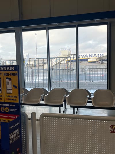 The scene depicts an indoor airport waiting area at Hahn Airport, near Lautzenhausen, Germany, viewed through large windows facing the tarmac under an overcast sky. In the immediate foreground to the left, a tall blue and yellow Ryanair sign details baggage policies, including "NON-PRIOR" boarding information, illustrations of "SMALL BAG" and "CABIN BAG" sizes, a QR code, and text indicating "BAG MUST FIT WITHIN THE BLUE & YELLOW LINE." To the right of the sign, four empty light-grey molded plastic chairs are arranged in a row, fixed to a black metal frame. The lower right foreground shows a white perforated metal barrier, possibly a queuing divider, with a small red and white sticker partially visible.
Beyond the windows, the airport tarmac is visible. Metal crowd control barriers are positioned directly outside. A pedestrian crosswalk with white stripes is marked on the asphalt. In the midground, a set of white air stairs with a blue handrail, marked "hahnairport" on its side, is positioned against a Ryanair aircraft. The aircraft, with its distinctive blue and yellow livery and "RYANAIR" branding, is parked on the tarmac. A tall light pole stands in the distance. The ground beyond the immediate tarmac appears to have light snow or frost. No individuals are present in the image.