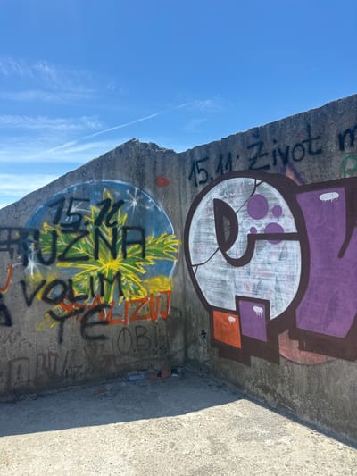 The image captures a section of a concrete structure, possibly a wall or building remnant, in the city of Gornji Kokoti, Montenegro. The structure is covered in graffiti. On the left, a circular design is painted in blue and yellow, with a green, star-shaped motif in the center. Black lettering, appearing to be "15.11. PUZNA VOLIM" is visible within and around this design. To the right, a large, stylized character is painted in purple and white, with brown outlines. The character has a circle with several smaller purple circles inside, and cracks are visible in the white paint. Some smaller graffiti in orange and red is present on the lower part of the wall. The background features a clear blue sky with scattered wispy clouds and a faint contrail from an aircraft. The ground in the foreground is a rough, concrete surface, with some debris, including bricks and a plastic bottle, visible near the base of the wall. The lighting suggests it is daytime, with a distinct shadow cast on the ground from the wall.
