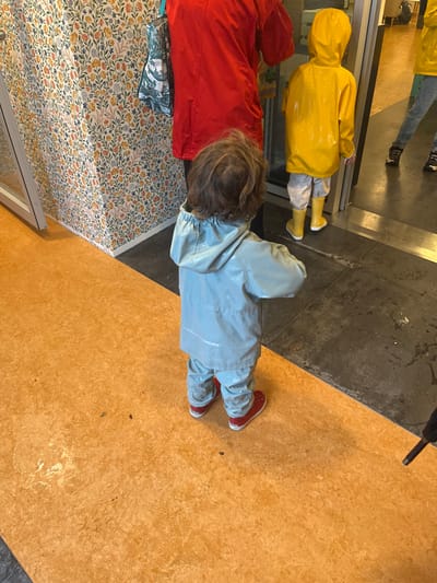 The image depicts an indoor scene in Vevey, Switzerland, featuring three individuals and a doorway. In the foreground, a young child, viewed from behind, stands centrally on an orange-ochre textured floor. This child wears a shiny, light blue two-piece waterproof suit with a hood and red, low-top shoes. To the left, a wall is covered in light-colored wallpaper with a dense floral pattern of small orange, blue, and green motifs. To the right of the foreground child, an adult figure is partially visible, wearing a red top and carrying a green and white patterned tote bag over their left shoulder. Adjacent to the adult, a second child, also viewed from behind, wears a yellow hooded rain jacket, light-colored trousers, and yellow rain boots. This child faces a dark-framed doorway with a dark threshold leading into another interior space. The floor immediately surrounding the doorway is dark grey/black and appears damp or reflective in areas. A reflection in the doorway shows the lower legs and black-shoed feet of a third individual. A black umbrella handle is partially visible at the bottom right corner of the frame. The scene suggests a transition point, likely an entrance or exit, possibly for individuals dressed for wet weather.