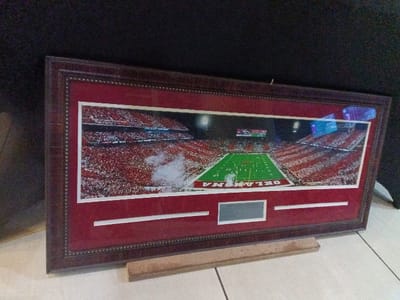 A framed panorama photograph depicts a packed football stadium at night. The image shows the field, stands filled with fans in red attire, and the stadium lights. "OKLAHOMA" is printed on the field. The frame is dark wood with a red matte. Below the photograph are two horizontal white bars and a small, gray rectangular plaque. The framed image is leaning against a dark background and resting on a tiled surface with a small wooden block underneath.