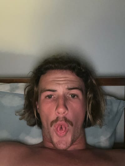 The image depicts a close-up, chest-up view of a single Caucasian male, approximately 20-35 years old, in an indoor setting identified as a location in Coalcliff, Australia. The man has medium-length wavy brown hair, a light brown mustache, and light-colored eyes. He is looking directly at the camera with a direct gaze, his mouth open in an O-shape, and his tongue curled longitudinally. His bare shoulders are visible, suggesting he is shirtless. He is positioned against a light blue pillow, which rests against a darker wooden headboard or frame. The background behind the headboard is a light, plain wall, possibly light grey or cream. The lighting is subdued, suggesting an indoor evening or low-light environment.