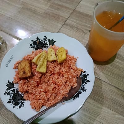 The scene, located in Ketowan, Indonesia, depicts a meal served on a light brown, wood-grain patterned surface. A white ceramic plate with scalloped edges and a black floral pattern along the rim, bearing the inverted name "JAMILIA," holds a portion of red-tinted fried rice. Five pieces of fried tofu are arranged on top of the rice; one larger, rectangular piece is golden brown, and four smaller, pale yellow, triangular pieces are also present. A silver-colored metal spoon rests on the plate, partially submerged in the rice. To the right of the plate, a clear plastic cup contains an orange-colored beverage with a blue straw inserted. There are no people visible. The objects are static, indicating the meal is prepared and presented, ready for consumption.