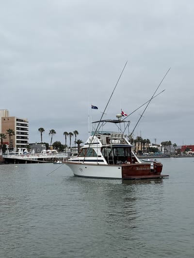 In the waters of La Punta, Peru, a white and wooden fishing boat named "MOOLOOLABA CALLAO" is anchored. The boat, equipped with multiple fishing rods, is positioned in the foreground, while in the background, a long pier lined with palm trees extends into the distance. Beyond the pier, a multi-story building with balconies and a coastal town with red-roofed buildings are visible. The sky is overcast, and the water is calm.