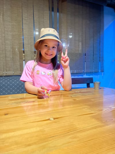 In this media file, a young girl, likely around 5-7 years old, is the central subject. She is wearing a pink t-shirt with an all-over print of cartoon characters, including Hello Kitty and other cute figures, and a straw hat with a blue band. Her right hand is raised, displaying a peace sign with brightly colored nails, and she holds a small toy in her left hand. She has a wide, happy smile, suggesting a joyful mood.
The girl is seated at a wooden table, which occupies a significant portion of the frame. The background features vertical blinds or screens with a textured pattern, and beyond that, a blue illuminated wall, possibly indicating an indoor setting with artificial lighting. A small metal bucket is visible attached to a dark support structure in the background. The overall lighting suggests it could be daytime, but the interior setting and controlled lighting make it difficult to be certain. The scene appears casual and possibly recreational, perhaps at a cafe or a play area. There is no visible text in the image. The orientation of the image is such that the subjects appear sideways.