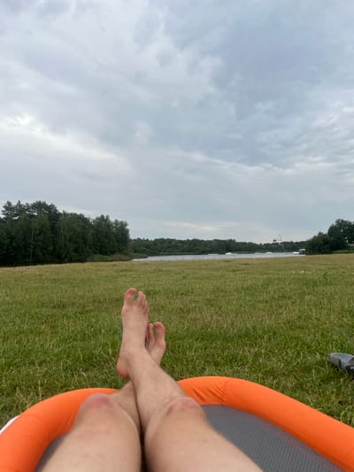 This is a first-person perspective shot of someone relaxing on an orange inflatable object on a grassy field. Their bare feet and lower legs are visible in the foreground, crossed at the ankles. The person appears to be at a water sports park, possibly in or near Enschede, Netherlands. In the background, a body of water is visible, with trees lining the far bank. Several water sports features, such as ramps and a cable tow system, are present on the lake. The sky is overcast and cloudy.