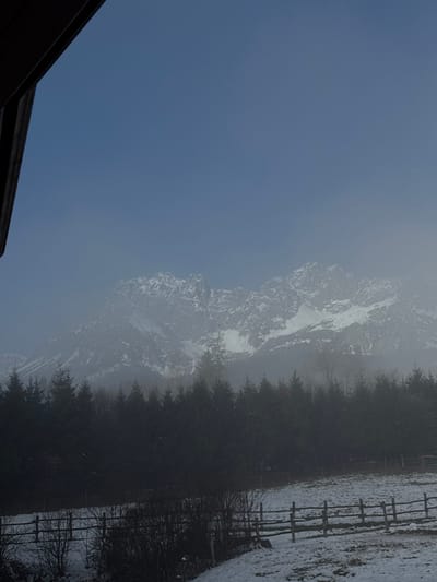 The image depicts a winter landscape in Going am Wilden Kaiser, Austria. In the foreground, snow-covered ground is visible, traversed by a wooden post-and-rail fence. Several dark, bare shrubs or small trees grow along the fence line. A dense band of dark green conifer trees forms a midground forest, partially obscured by a light haze or fog. In the background, towering snow-capped mountains, consistent with the Wilder Kaiser range, rise into a sky that transitions from hazy light blue at the mountain peaks to a clearer, darker blue overhead. The upper left corner of the frame reveals a dark brown wooden architectural element, likely an eave or beam, indicating the photograph was taken from under an overhang. No people or animals are present.