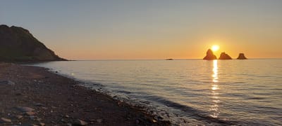 The image shows a serene coastal scene at sunrise or sunset. The foreground features a rocky beach with scattered stones. The sea stretches out towards the horizon, with gentle waves lapping at the shore. In the middle ground, three distinct rock formations rise from the water, silhouetted against the vibrant orange and yellow hues of the sky. The sun, partially obscured by one of the rock formations, casts a shimmering reflection on the water's surface. To the left, a green, sloping hill descends towards the sea, its texture suggesting vegetation. The sky transitions from a pale blue at the top to a warm gradient of orange and yellow near the horizon. The location is identified as Aleksandrovsk-Sakhalinskiy, Russia.