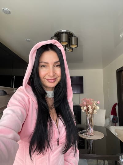 An indoor scene in a residential location within Syasstroy, Russia, features a woman taking a selfie. The woman, positioned in the foreground and center, has long, straight dark hair and a light smile. She wears a light pink, zippered velour or velvet hooded jacket, and a multi-strand silver-colored choker necklace. A black garment strap is visible beneath the jacket's partially unzipped front.
The background shows a contemporary interior. The ceiling is white, with recessed lighting and a central chandelier featuring five dark cylindrical shades and clear dangling elements. To the left, dark cabinetry or furniture is visible. In the mid-ground, a dark, reflective rectangular table is present, holding a tall clear glass vase with dried light pink and white flowers, a light beige placemat, a pair of dark-framed eyeglasses, and a remote control. A flat-screen television is mounted on the wall in the center background. To the right of the table, two light-colored chairs are partially visible. Further right, a dark-framed window or doorway is next to a wall on which a red gnome decoration with a pointed hat is displayed.