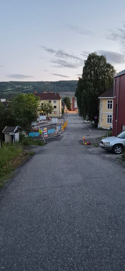 The image depicts a street in Lillehammer, Norway, under an overcast sky. The road is paved and slopes upwards into the distance, lined with residential buildings and trees. Construction barriers, adorned with blue tarps and yellow and white cones, are visible along the left side of the road, indicating ongoing work. A small, shed-like structure with a blue door and white trim is situated to the left of the construction site. On the right side of the road, a silver SUV is parked. The background features a densely wooded hillside with scattered houses, characteristic of the Norwegian landscape. The overall impression is a quiet, residential street in a hilly area, with some disruption due to construction.