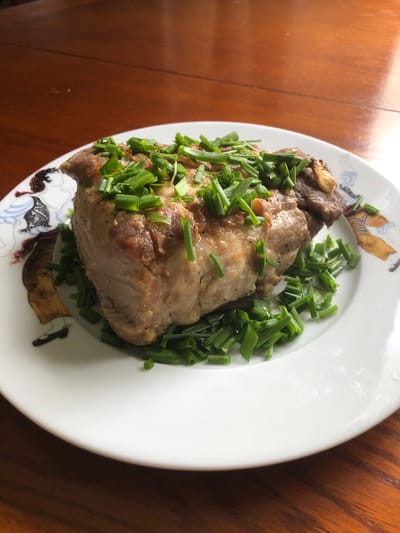 A close-up, overhead shot captures a white plate filled with a savory dish. The main subject is a large piece of cooked meat, possibly pork or chicken, seasoned and browned to a rich color, topped generously with finely chopped green chives. The meat has a rustic appearance, with visible texture and some darker, caramelized edges. The chives are fresh and vibrant green, scattered both on top of the meat and around the edges of the plate. The plate itself is decorated with a subtle pattern featuring stylized figures and abstract shapes in muted colors, visible along the rim.

The plate rests on a warm, brown wooden surface, likely a table, with natural light casting gentle shadows and highlights on both the food and the background, suggesting it's daytime. The setting appears to be indoors, possibly a home dining area. No people are visible in the image. The food is the sole focus, presented simply and invitingly. There is no visible text in the image.