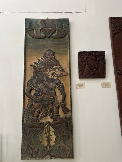 This media file captures a display of carved artworks on a white wall, likely in a museum or gallery setting in Cirebon, Indonesia. The primary focus is on two dark wooden relief carvings.
The larger, lower carving is a horizontal panel featuring a dynamic, stylized depiction of a figure that appears to be a mythological character or warrior. The figure has an elaborate headdress, intense facial features with wide eyes and bared teeth, and is rendered in a dramatic pose with limbs outstretched. The carving is intricate, showcasing detailed patterns in the clothing, armor, and surrounding decorative elements. The background of this panel has a textured, earthy tone with hints of green and brown pigment, suggesting a weathered or aged appearance. The overall style evokes traditional Indonesian art forms, possibly influenced by Wayang puppetry.
Above this larger piece, a smaller, square wooden carving is displayed. It also features a detailed relief, though the subject matter is less distinct at this resolution. Two small, rectangular paper labels are affixed to the wall next to this smaller carving. The visible text on these labels reads: "UKIRAN WAYANG / PATIMUR" and "UKIRAN WAYANG / R KUMBA KARNA", with "1720 M" also visible on both. These labels suggest that both carvings are "Wayang carvings" and may be dated to 1720.
The setting appears to be an indoor environment with bright, even lighting. The wall is painted white, and there are some subtle signs of age or imperfection on the wall's surface. The arrangement suggests a curated exhibition of cultural artifacts.