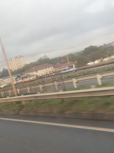 The image depicts a multi-lane highway scene in Theta ward, Kenya, under an overcast sky. In the immediate foreground, a dark asphalt road surface with a faded yellow line on the left is visible. A metal guardrail, composed of multiple horizontal rails and concrete posts, separates the viewer's current lane from the adjacent traffic lanes. Motion blur suggests movement from the viewer's perspective.

In the midground, traffic is present on the highway. An orange and white public transport bus (possibly a matatu) with "C.P.O" visible on its side is either stationary or moving slowly on an outer lane. Several indistinct dark figures, presumably people, are visible near the bus. A light-colored sedan is positioned on a lane closer to the viewer, and a white van or smaller bus is visible behind the orange and white bus.

The background features urban infrastructure and natural elements. On the left, a tall, multi-story rectangular building, light-colored with numerous windows, stands prominently. Closer to the highway, a lower-rise building with a reddish-brown tiled roof and white walls is visible. Extensive green vegetation, including numerous trees, is spread across the background, interspersed with other distant buildings. A slender, light-colored pole is partially visible on the far left. The sky is uniformly light grey, indicating overcast conditions.