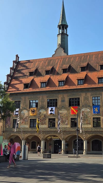 A multi-story building with an orange tiled roof and a tall green spire is visible under a clear blue sky in Ulm, Germany. The building's facade is adorned with murals and flags. There are arched walkways on the ground level. In the foreground, a woman in a pink suit walks on a paved area. Other people are seated on a bench to the left, and one person walks towards the building's entrance. Several flags are displayed, including one yellow flag with a black eagle and another blue flag with a yellow symbol. The building appears to be a historic structure, possibly a town hall or a prominent landmark.