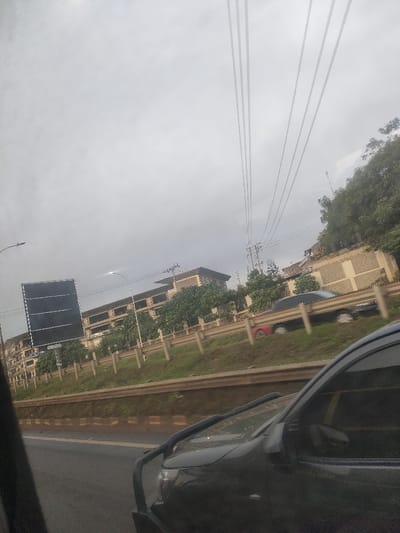 The image captures an overcast daytime scene in Gatongora ward, Kenya, observed from within a vehicle. In the immediate foreground, the front-right portion of a dark-colored vehicle, featuring a bull bar, is partially visible. A multi-lane roadway extends from the lower-left, bordered by a concrete barrier that separates it from a grassy median. A metal guardrail runs horizontally across the midground. Beyond this guardrail, a row of green trees partially obscures multiple structures. A prominent multi-story building with a light-colored facade and a dark roof is centrally located behind the foliage. To the left of this building, a large, dark, blank billboard stands near a street light pole. Two additional vehicles, a red car and a darker sedan, are partially visible on a road or parking area behind the guardrail. The sky is uniformly gray and overcast, with several parallel power lines traversing the upper frame from left to right, supported by a visible utility pole on the right. No human figures are discernible.