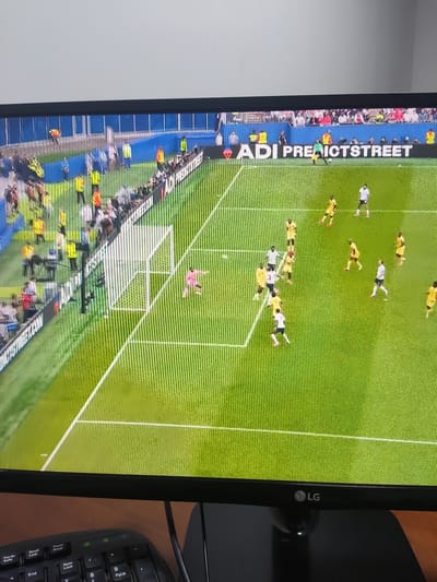 The image shows a screen displaying a soccer match. The field is green with white lines. There are several players on the field wearing yellow and white jerseys. In the background, there are spectators and advertising banners. The bottom of the image shows a portion of a computer monitor with an LG logo and a keyboard. The scene does not depict any specific location in the city of Egizbulak, Uzbekistan.