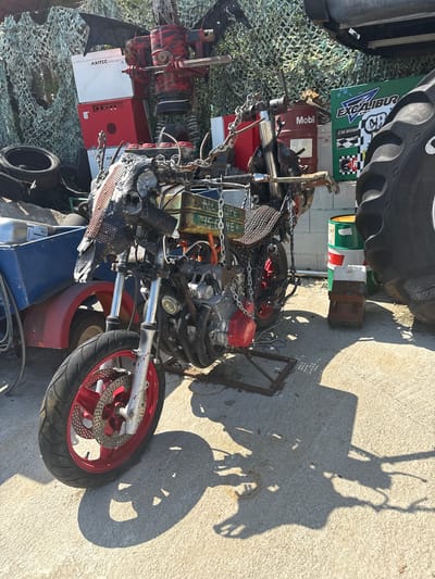 The image shows a cluttered outdoor scene, possibly in the city of Općina Funtana, Croatia, though there are no geographic markers present. A disassembled or custom-built motorcycle, comprised of various parts and materials, is central. Components visible include tires, an engine, a red fuel tank, chain, and metal scrap. Several unrelated objects are adjacent to the motorcycle: a large tractor tire, a Mobil oil barrel, an Axitec solar storage unit, and miscellaneous metal containers. The area is paved with concrete, and the scene is partially covered with camouflage netting. Strong sunlight casts distinct shadows of the motorcycle and surrounding objects onto the pavement. There are no people visible.