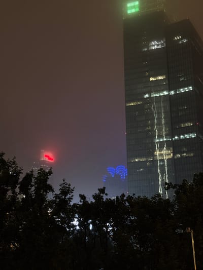 This image captures a night scene of modern city architecture in Jing'an District, China. The primary subjects are two tall, contemporary skyscrapers, partially obscured by fog or mist, which lends a dramatic and somewhat mysterious atmosphere to the scene. One skyscraper on the left has a facade of dark glass with rows of illuminated windows, reflecting light in horizontal patterns. A section of this building is lit with a bright green light.
Further to the right, a second skyscraper features vibrant blue, intricate light patterns, resembling a stylized crest or radiating design. Even further in the distance, a third building is visible, illuminated by a bright red neon sign that appears to spell out characters, though they are not clearly decipherable.
The foreground is dominated by the dark silhouettes of lush green trees, their leaves and branches forming a natural frame for the urban landscape. The night sky is dark, with the city lights and building illuminations providing the main sources of light. The fog creates a diffused glow, softening the harsh edges of the buildings and contributing to a sense of depth and scale. The overall mood is one of urban grandeur and modern spectacle, with a touch of atmospheric mystery due to the fog. The time of day is clearly night.