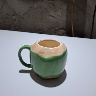 This media file contains a close-up, slightly angled shot of a ceramic mug. The mug is predominantly green, with a beige or light brown band around the opening and a matching handle. The interior of the mug is a warm, glossy yellow. The mug is positioned on a white surface, casting a distinct shadow to its right.
In the background, to the left of the white surface, is a textured, dark, and uneven surface that appears to be a wall or floor. A thin, orange and yellow wire runs horizontally across this dark background, suggesting an indoor or possibly outdoor workspace.
The lighting appears to be artificial, creating sharp shadows and highlighting the glossy finish of the mug. No people are visible in the image, and no text is present. The overall impression is that of a studio shot showcasing a handmade ceramic object. The location context of Reudeuep, Indonesia, is noted but does not provide explicit visual cues within the image itself.