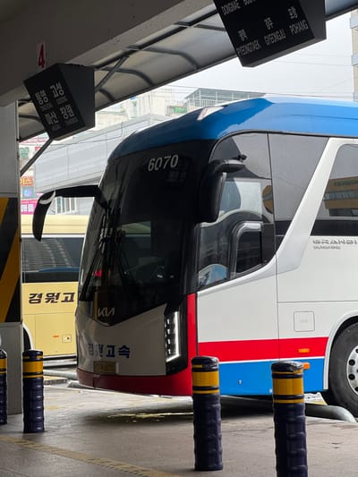 This is a photograph taken in Geoje-si, South Korea, depicting a parked bus at what appears to be a bus station or terminal. The bus is predominantly blue and white with red accents. On the side of the bus, the text "GRANBI SUNSHINE" is visible. The bus also has "6070" written on its front. There are black and yellow bollards in the foreground, suggesting a designated parking area.
To the left of the bus, a black signpost displays route information in both Korean and English. Text on this sign includes place names such as "DONGNAE," "PYEONGTAEK," "GYEONGJU," and "POHANG," as well as "TONGYEONG," "GOSEONG," "MASAN," and "CHANGWON." This indicates that the bus serves routes to these locations.
In the background, a yellow bus is partially visible, and architectural elements of a building or structure are present. The lighting suggests it is daytime, and the ground appears to be slightly wet, hinting at recent rain or damp conditions. The overall scene conveys a sense of urban transit infrastructure. The focus is on the bus and its associated signage, highlighting its role in regional transportation.