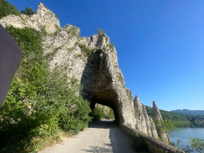 The image shows a road passing under a large natural rock arch with a clear blue sky above. The road is made of gravel and a stone wall runs along the right side. Lush green trees and vegetation are visible on the left, and the rock formations on the right appear jagged and pointed. In the background, a body of water is visible, reflecting the sky, with rolling green hills and mountains in the distance. This scene is a natural landmark located in Asparuhovo, Bulgaria, known as the "Stone Forest" or "Kamenni Gorat." There are no people visible in the image.