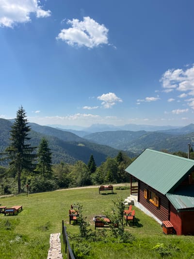 The image depicts a scenic view from Katun Kobil do, Montenegro, featuring a wooden cabin with a green roof situated on a grassy slope overlooking a vast expanse of rolling, forested mountains under a clear blue sky dotted with fluffy white clouds. In the foreground, a pathway made of stone slabs leads down from the viewer's perspective. The cabin's exterior is made of dark red wood with orange-tinted windows and shutters. Several wooden benches and tables are arranged on the lawn around the cabin, some with small fire pits. A tall evergreen tree stands prominently to the left of the cabin. The distant mountains fade into layers of blue and gray, indicating a great distance. The environment is natural and undeveloped, suggesting a remote mountain retreat. There are no people visible in the image.