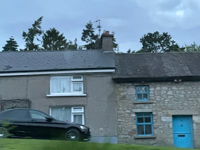 The image shows a row of two houses in an unclear location in Ireland. The house on the left has a gray exterior and is taller, featuring two windows on the upper floor and two on the lower floor, all with white frames and white curtains. A black car is parked in front of this house, positioned on a green lawn. The house on the right is constructed from stone and appears older, with two windows on the upper floor and one on the lower floor, all painted bright blue. A bright blue door is also visible on the right side of this stone house, with a rectangular cutout near the bottom. Trees are visible behind the houses, and the sky is overcast. The image appears to have been taken during daylight hours, possibly late afternoon or evening, given the subdued lighting.