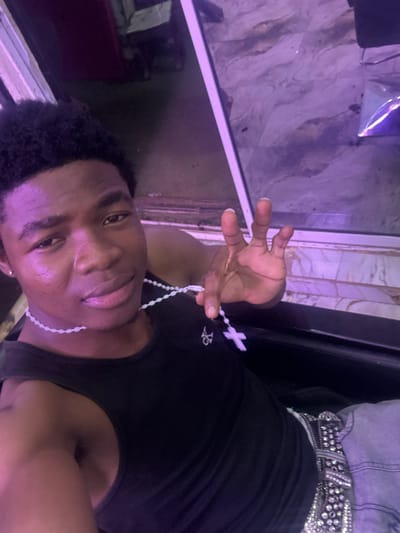A portrait of a person with dark skin and short black hair, wearing a black tank top, a shiny silver belt, and a white beaded necklace. The person is holding the necklace up with their right hand. The background is a blurry combination of purple and white hues, suggesting indoor lighting and possibly a tiled or patterned surface. The image does not provide enough detail to determine its location in Ogwashi-Uku, Nigeria.