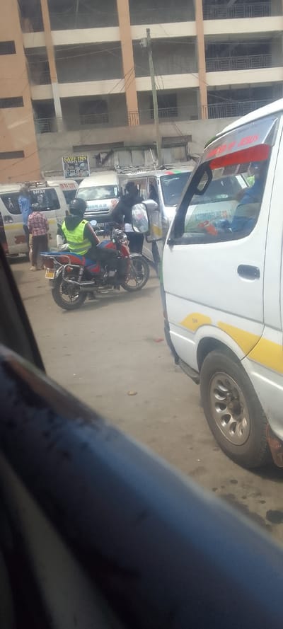 The image depicts an urban street scene in Naivasha, Kenya. In the foreground, the lower left quadrant is partially obscured by the dark interior of a vehicle. A white van (matatu) with yellow lower body accents and a "Turbo" inscription is positioned on the right, displaying a red sun visor with "POWERED BY JESUS". A person in a blue shirt occupies the passenger seat.

In the middle ground, a red and blue motorcycle with a partially visible license plate "KMCD 671G" is stationary. The rider wears a black helmet and a bright yellow-green reflective vest. To the left of the motorcycle, a male individual in a red and white checkered shirt and light-colored pants stands near a white van labeled "NEXT". Further back, another person in dark clothing appears to be interacting with a white van that bears a "KIKIMA MILLING CO. LTD" sign.

The background consists of a multi-story building, light brown in color, featuring multiple windows and balconies with dark railings. Below this building, a "SAVE INN CHIPS & FISH" sign is visible. A wooden utility pole with overhead wires stands centrally in the background. The ground is an unpaved or damaged road surface. The overall scene suggests a busy commercial street with multiple vehicles and pedestrians.