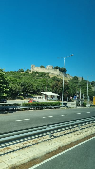 The image displays a clear, bright blue sky above a verdant hillside topped with a stone fortress. A paved road runs horizontally across the foreground, with a metal guardrail separating it from a sidewalk and a strip of vegetation. Several vehicles are visible on the road, including a red car and a dark blue car. Adjacent to the road, a small, white, open-air structure with a roof is situated on the hillside, possibly a bus stop or a small kiosk. Streetlights are positioned along the road, and the fortress on the hilltop features multiple turrets and crenellated walls, suggesting historical significance. The environment appears to be a sunny day, with ample greenery surrounding the fortress and the roadside. The scene is located in Neos Panteleimonas, Greece, near Platamonas Castle.