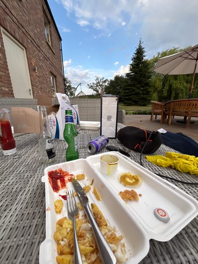 A messy outdoor table setting displays remnants of a meal and beverages. A polystyrene takeaway container holds leftover fish and chips with a knife and fork. Various empty bottles, including beer and a ketchup bottle, are scattered around. An open purple can sits near a cardboard box. In the background, glimpses of outdoor furniture, greenery, a brick building, and a cloudy sky are visible.