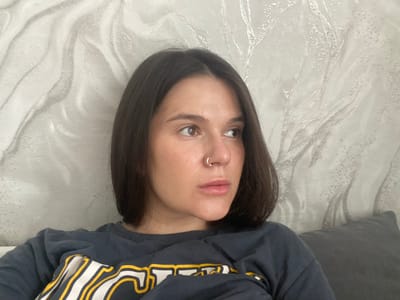 A young woman with dark brown hair and a nose ring is pictured. She is wearing a dark grey sweatshirt with yellow and white lettering. The woman is looking to her right, and her face is partially visible. She is seated on a couch, with a grey pillow behind her. The wall behind the couch has a textured white wallpaper with a subtle silver pattern. The lighting in the room is soft and diffused, casting gentle shadows. The scene appears to be an indoor residential setting, possibly a living room or bedroom. There is no discernible information in the image to suggest it is located in Romny, Ukraine.