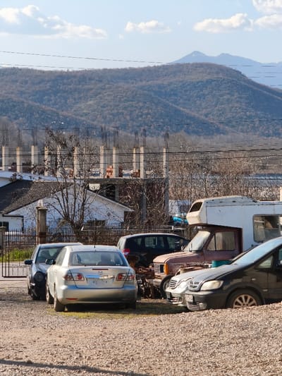 The image displays an outdoor scene in Mrzenci, North Macedonia, under a bright, partly cloudy sky. The foreground consists of light-colored gravel. Several vehicles are parked on this gravel surface. From left to right, a dark sedan is partially visible behind a silver Mazda 6 sedan, which is positioned with its rear facing the viewer. To the right of the Mazda, a dark hatchback is visible behind a rust-colored pickup truck fitted with a white camper shell. Further right, a dark minivan is partially visible. In the midground, behind the vehicles, a white single-story building with a dark roof is seen on the left, fronted by a metal gate and a concrete pillar. To its right and further back, a multi-story concrete structure under construction is visible, featuring numerous exposed concrete pillars with rebar extending upwards and stacks of red bricks on a lower level. Bare deciduous trees and shrubs are scattered throughout the midground. The background features a range of hills or mountains covered with sparse, bare vegetation. Utility wires traverse the mid-upper portion of the image horizontally. No individuals are present in the scene.