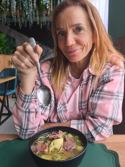 A woman with reddish-blonde hair is sitting at a table, holding a spoon in her right hand and looking at the camera with a slight smile. She is wearing a pink t-shirt under a pink plaid shirt. In front of her is a black bowl filled with soup, which contains noodles, meat, and potatoes, resting on a green scalloped placemat on a wooden table. The background features a blurred restaurant or cafe setting with green plants, marble walls, and blue chairs. The location is Semenovka, Kyrgyzstan.