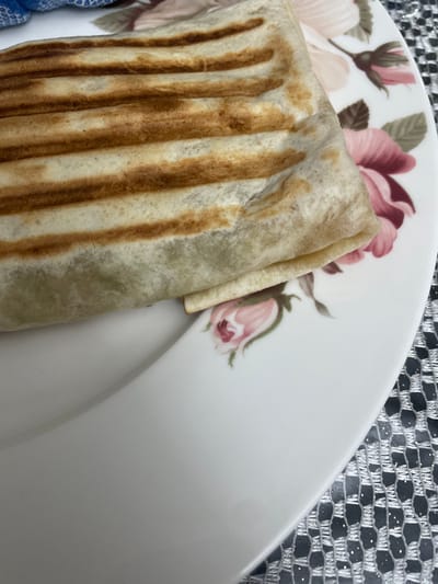 A folded wrap with grilled markings sits on a plate adorned with floral patterns. The wrap is light in color, with dark brown grill lines. Part of the plate features a design of pink and burgundy flowers with green leaves. In the background, a textured silver surface is visible.