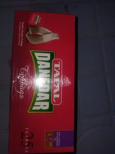 A close-up, horizontal, medium shot captures a red cardboard box of Tapal Danedar tea bags. The box is sealed in clear plastic. The brand name "DANEDAR" is prominently displayed in large white, green, and yellow lettering across the center of the box, with "TAPAL" appearing in a vertical red oval to its right. The phrase "Double Chamber Teabags for quicker infusion of tea" is printed in white text on the left side of the box. Towards the bottom left, the number "25" is printed in large black font, with "TEABAGS" printed vertically next to it. A small purple and gold seal with "TAPAL TEA" is visible on the lower right. The background consists of dark gray, textured floor tiles. The image is likely taken indoors, as indicated by the artificial lighting causing glare on the plastic wrapping and the tiled floor. The location is identified as Haveli Jhangranwali, Pakistan.