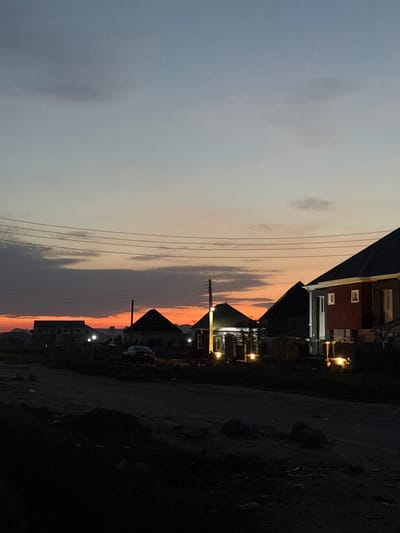 The image depicts a twilight scene in Angwan Sayawa, Nigeria, characterized by a vibrant sunset and residential architecture. The sky transitions from a light blue-grey at the top to a rich orange and red hue on the horizon, partially obscured by dark, streaky clouds in the mid-sky. Distant hills are silhouetted against the bright horizon on the left.

A series of power lines span across the middle ground, supported by utility poles, one of which is prominent on the center-right. Below the power lines, several houses are visible. On the far left, a multi-story building is silhouetted. Towards the center-left, a house with a dark, sloped roof stands, accompanied by a bright white exterior light. A dark sedan is parked on the unpaved road in front of another house with a dark, pyramidal roof, which is illuminated by multiple warm yellow lights. On the far right, a two-story house with a reddish-brown facade, white window frames, and a dark, sloped roof is brightly lit with warm yellow exterior lights at both ground and upper levels.

The foreground consists of a dark, unpaved dirt road with uneven terrain, devoid of clear markings or pavement. The ground appears dry and dusty, with some shadowy vegetation or debris along the immediate bottom edge. No people are discernible in the scene.
