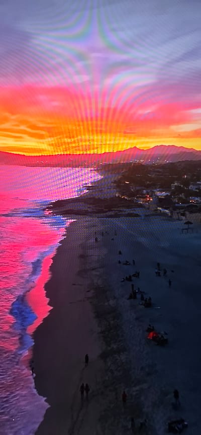The image depicts a vibrant sunset over a coastal landscape. The sky is a dramatic gradient of fiery orange and bright pink, transitioning to a soft purple and blue at the top. Rays of light emanate from the horizon, creating a luminous effect. Below the sky, distant mountains are silhouetted against the colorful backdrop, with faint lights suggesting a town or city.

The foreground features a beach scene. The ocean's edge is illuminated by the sunset, reflecting the pink and purple hues. Waves gently lap the shore. The beach itself is sparsely populated with a few individuals and small groups scattered across the sand. Some people are walking along the shoreline, while others are gathered in small groups, possibly relaxing or socializing. There are also indications of beach equipment, such as umbrellas or tents, in a few areas. The overall atmosphere is one of tranquility and natural beauty, enhanced by the dramatic colors of the sunset. The image does not provide any information that can place the location in Lippstadt, Germany.