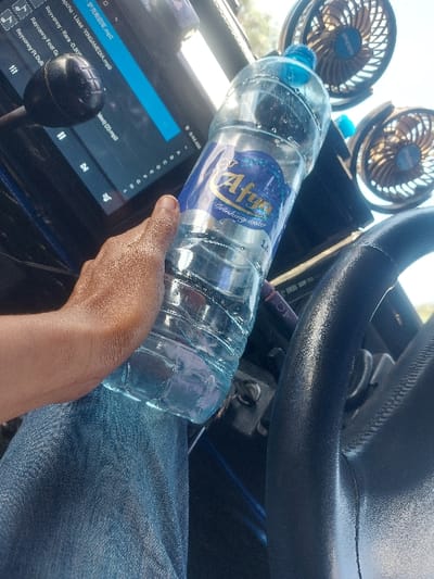 Inside a vehicle located in Kimamba, Tanzania, a person with dark skin is depicted from the mid-torso down. Their right hand is holding a 1.5-liter clear plastic bottle of Afya Drinking water, which rests against their blue denim-clad thigh. The bottle contains water and exhibits condensation on its exterior. To the left of the person's hand, a digital screen displays a music player interface, showing song titles including "Rayvanny - Rara" and "Rayvanny Feat G...", with the time "8:44 am" visible. A black gear stick is positioned above the screen. A section of a black steering wheel is visible on the right side of the frame. Two small, dark-colored portable fans with blue-accented blades are mounted on an upper surface within the vehicle's interior. The scene is illuminated by natural daylight from outside the vehicle.
