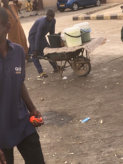 The image depicts an outdoor street scene in Unguwar Fatika, Nigeria. In the midground, a dark-skinned male wearing a blue long-sleeved tunic, matching trousers, and dark sandals with yellow branding is positioned behind a rusty, single-wheeled wheelbarrow. He is bent forward, pushing the wheelbarrow and looking towards the viewer. The wheelbarrow's green basin contains a black bucket, a small clear container holding light-colored solid items, and two lidded clear plastic buckets. One clear bucket holds chopped white produce, and the other holds chopped green produce. A light-colored fabric is draped over the wheelbarrow's bed.

In the foreground, partially visible on the left, a dark-skinned male wears a navy blue short-sleeved polo shirt with a "GOD IS G..." logo and black trousers. He holds an orange and black object, consistent with a measuring tape, in his right hand. To the left in the background, a male in a light brown patterned garment is partially visible. Further back, a blue hatchback car with the license plate "YD10 VGP" is parked. Behind the car, a low curb is painted with yellow and black stripes. Several indistinct male figures are seated or crouching on a raised platform in the far background. The ground is a grey, uneven surface, likely concrete or asphalt, marked with dark liquid stains and scattered debris, including organic matter and plastic wrappers.