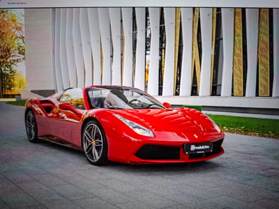 A bright red convertible Ferrari is parked on a paved pathway in front of a modern building with a white and gold facade. The car is the primary subject, facing towards the right of the frame. Its doors are open, revealing the interior. The building's facade is characterized by vertical white panels interspersed with gold-colored elements that appear to be textured or perforated. Green grass and fallen leaves are visible to the right of the pathway. The sky is not visible in the frame. The scene appears to be an outdoor setting in a city environment.