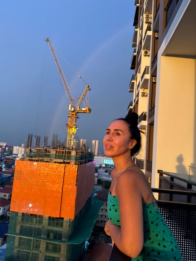 A woman with dark hair in a bun, wearing a green polka dot dress, stands on a balcony and looks up at a construction crane against a blue sky with a faint rainbow. She is in Khan Mean Chey, Cambodia. The building behind her is a modern apartment complex with multiple balconies and air conditioning units. In the background, a large building under construction is covered in orange netting. The lighting suggests it is either sunrise or sunset, casting a warm glow on the woman and the building.