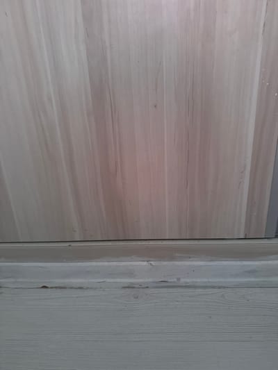 The image presents a close-up, static view of an interior architectural space, likely within a building in North Nicosia, Northern Cyprus / Cyprus. The composition is divided vertically between different textured surfaces.
On the left, a large panel displays a prominent light beige to pale brown horizontal wood-grain pattern. This surface, possibly a door or a wall panel, has a smooth finish with visible striations and a few minor, light-colored specks or marks scattered across it. Above this panel, a narrow, dark grey or metallic trim is visible along the top edge.
Separating the left panel from the right side is a slim, dark vertical line, suggesting a gap, seal, or thin frame element. To its immediate right is a light-colored, almost white, vertical surface that appears to be a door jamb or part of a frame. This section exhibits a rougher, possibly unfinished or weathered texture, with some irregular patches and dirt, suggesting it might be an unpainted or partially prepared surface like plaster or wood.
Further to the right, beyond this rougher frame, another vertical surface is visible. This material clearly displays a vertical wood grain pattern and is uniformly light-colored, possibly painted or stained in a pale grey or off-white tone, offering a textural contrast to the horizontal grain on the far left.
The lighting is consistent with indoor conditions, and no time of day, weather, or human presence can be inferred. No text is visible in the frame. The scene captures an interplay of utilitarian textures and muted colors, possibly indicating an area under construction, renovation, or a functional space.