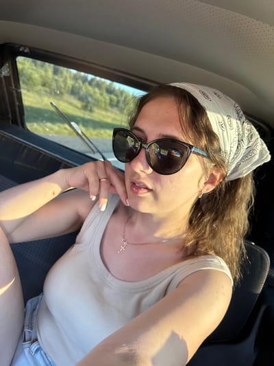 A young woman with light brown hair tied back with a white bandana is captured in a selfie taken from the backseat of a vehicle. She is wearing dark sunglasses, a light beige tank top, and denim shorts. A delicate gold necklace with a cross pendant is visible around her neck. The setting sun casts a warm glow on her skin. The background shows a green, tree-lined landscape viewed through the car window. The image appears to have been taken during daylight hours, with the lighting suggesting it could be late afternoon or early evening. There is no specific indication of the city of Yurkovo, Russia, in the image.