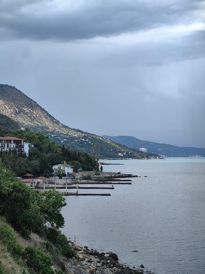 A wide shot captures a coastal view with a mountain on the left and the sea on the right. The sky is overcast with heavy clouds. On the mountainside, there are scattered buildings and dense green vegetation. In the foreground, trees and rocks line the shore. Several piers extend into the water, some with a few people visible and birds perched on them. The water is calm and appears to be a pale blue-grey. In the distance, along the coastline, more buildings are visible on the lower slopes of the mountains.