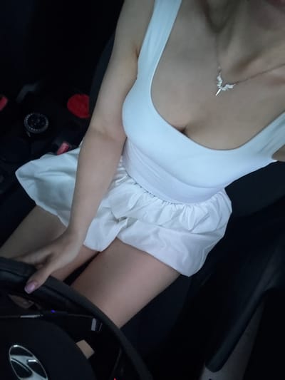 A person is sitting in the driver's seat of a car, hands on the steering wheel. The person is wearing a white tank top and a white skirt. A necklace with a winged pendant is visible around the person's neck. The car's interior is dark, with some red elements visible in the background, possibly part of the car's interior design or accessories. The scene appears to be in Petukhovo, Russia, based on the prompt.